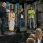 14.6.2026 Art:walk auf der Straße der Romantik und Revolution, der Bilker Straße. Manfred Hill begrüßt die Kinder und die Gäste zu Beginn des SingPause-Konzertes. Auf der Bühne die Singleiterinnen Maria Carreras und Clementine Schwander und der Singleiter Martin Lucaß