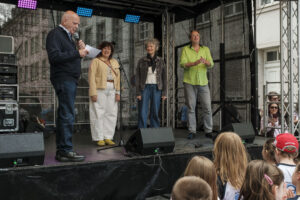 14.6.2026 Art:walk auf der Straße der Romantik und Revolution, der Bilker Straße. Manfred Hill begrüßt die Kinder und die Gäste zu Beginn des SingPause-Konzertes. Auf der Bühne die Singleiterinnen Maria Carreras und Clementine Schwander und der Singleiter Martin Lucaß