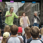 14.6.2026 Art:walk auf der Straße der Romantik und Revolution, der Bilker Straße. Auf der Bühne die Singleiterinnen Maria Carreras und Clementine Schwander und der Singleiter Martin Lucaß schwer im Einsatz.